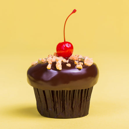 banana split cupcake with banana cake, marshmallow filling, and strawberry frosting, that has been dipped in ganache and finished with crushed peanuts and a maraschino cherry