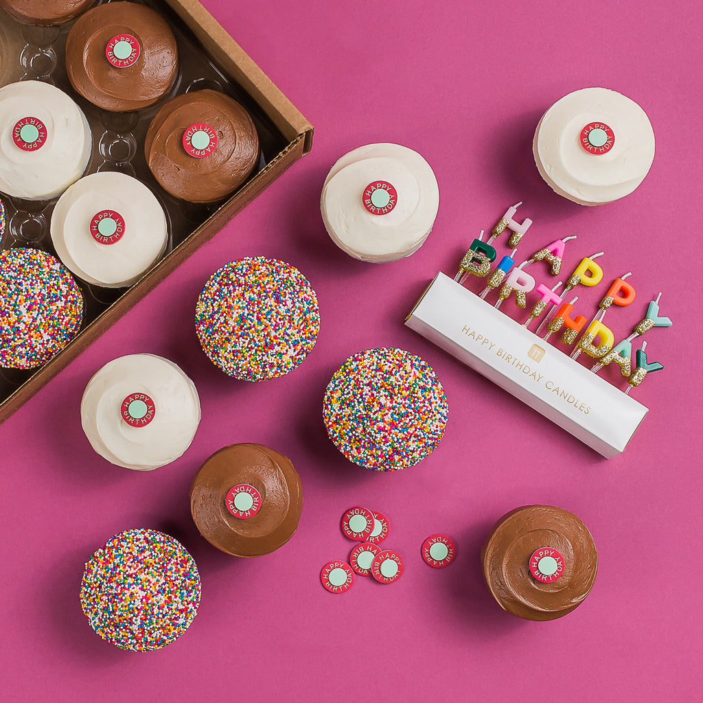 Birthday Box with 12 assorted cupcakes (red velvet, vanilla, dark chocolate, sprinkle), gold candles, decorations, and a birthday sticker.